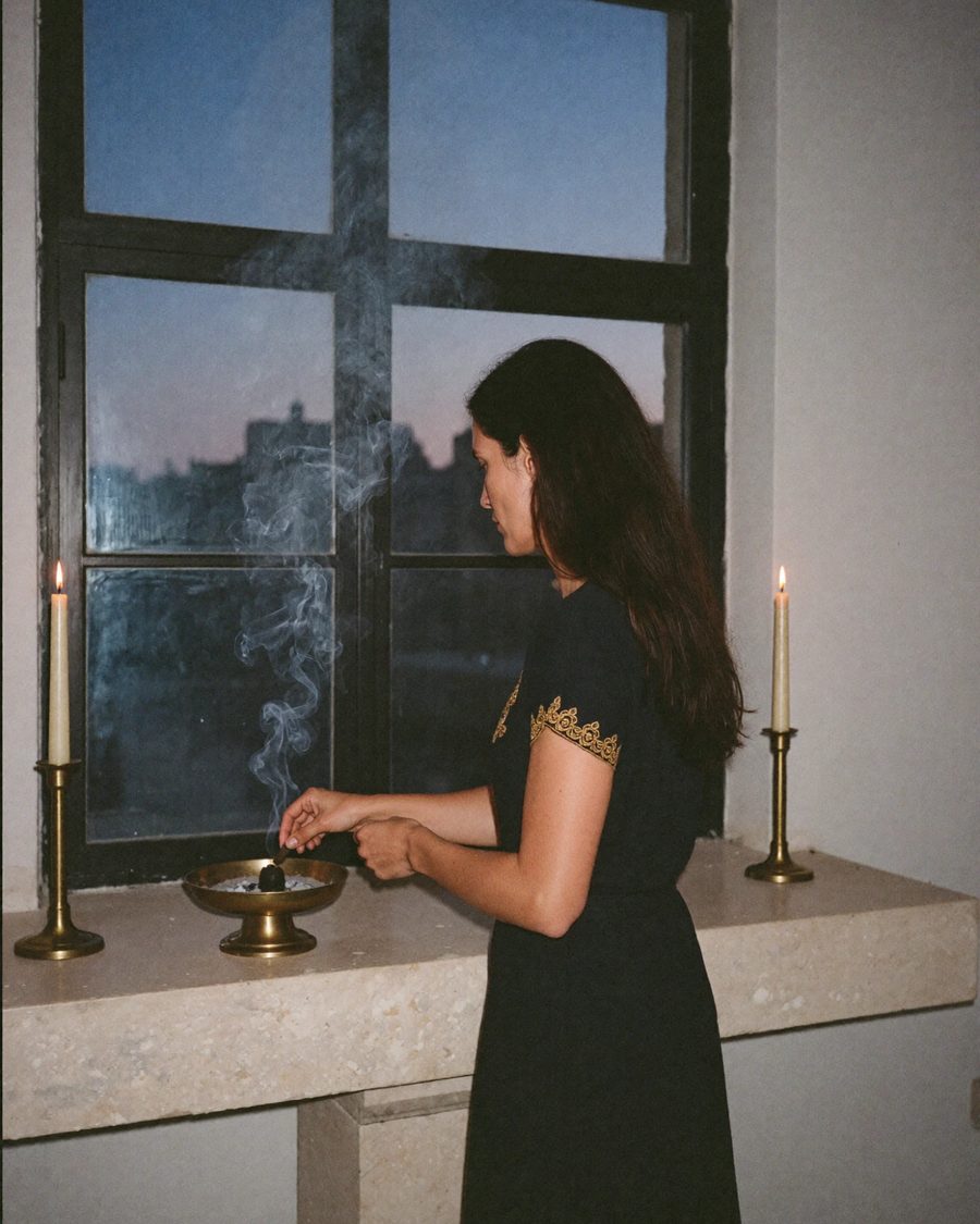 Amara as a younger woman, lighting incense between two candles at a window at dusk